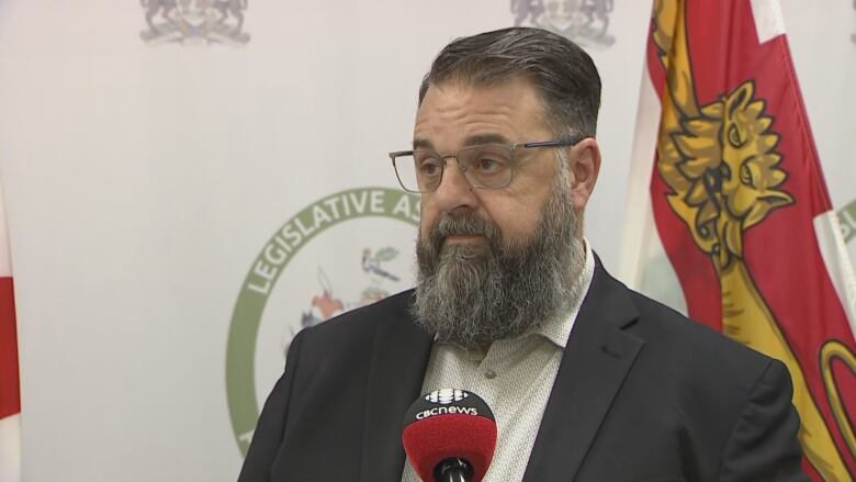 A man with a dark jacket, white shirt and beard taking questions in front of a P.E.I. Legislative Assembly backdrop.