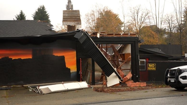 A building is seen being demolished. It is black and orange and there is a No Trespassing sign on a chain-link fence to the right of the structure. An RCMP SUV sits in front of the scene.