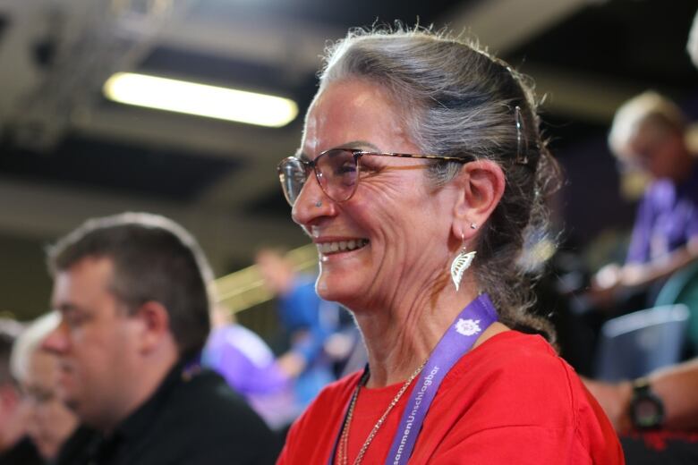 A white woman wearing a butterfly-wing necklace and a red t-shirt smiles in a crowd.