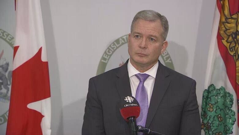 A man in a dark suit and purple tie stands in front of microphone taking questions from reporters.