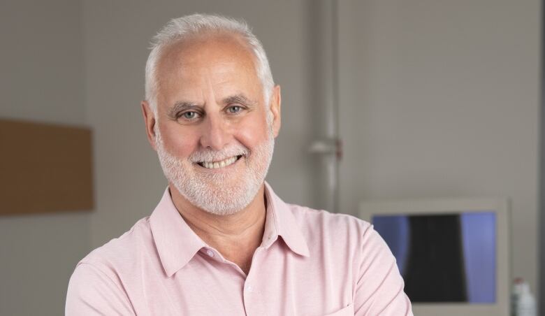 A man with white hair and beard smiles for a photo taken in a medical examination room with medical equipment seen in the background.