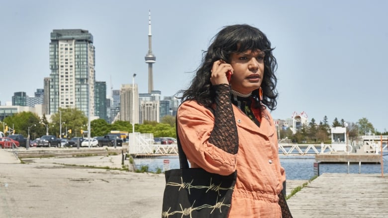 A person standing on a dock holds a cellphone to their ear. Behind them can be seen the CN Tower. 