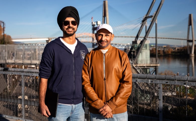 Two Indo-Canadian men smile for the camera, standing in the sunlight in front of a bridge over a river.