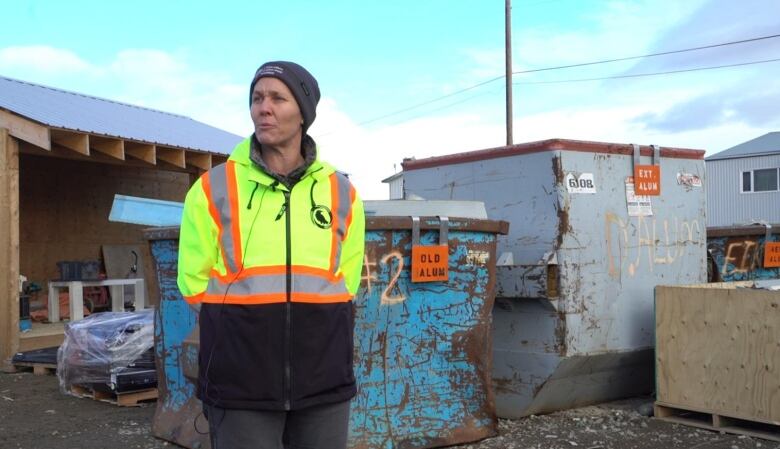 A woman in a bright safety jacket stands nears some dumpsters.