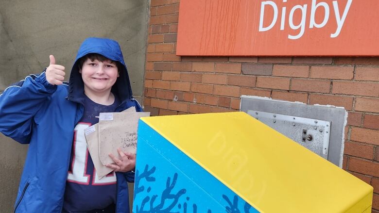 A young boy holds envelopes next to a Canada Post mailbox, smiles and gives a thumbs up.