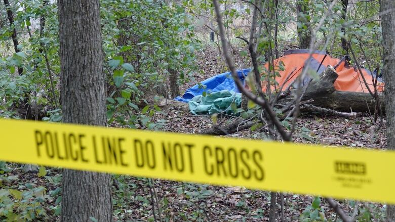 Yellow police tape is in the foreground of a photo showing what appears to be a collapsed tent and tarp in a wooded area.