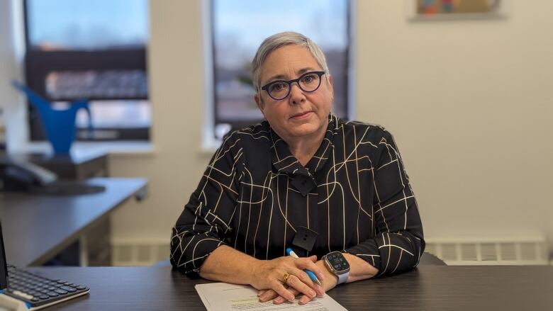 Woman with very short hair and glasses sits at a desk with a large window in the background. 