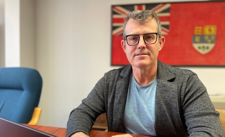 A middle-aged man with short hair and glasses sits at a desk inside an office. He looks at the camera. A flag hangs on the wall behind him.