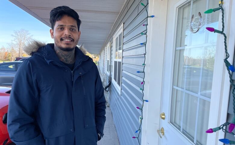 A man with short hair wears a winter coat and smiles outside of an apartment door. 