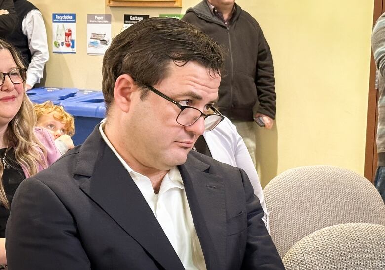 A man in a navy jacket and white shirt looks up over his glasses.