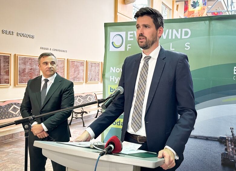 A tall man in a blue jacket, white shirt and plaid tie speaks at a microphone as another man listens.