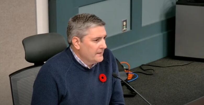 A person with short grey hair in a blue sweater sits in front of a microphone.