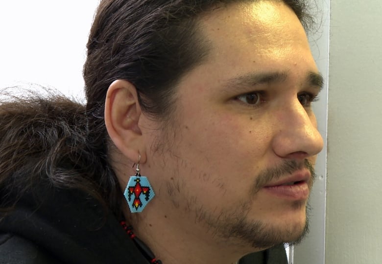 The face of a man with long dark hair and a wispy beard, wearing a beaded blue and red earring, is shown in profile.