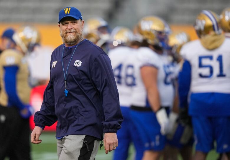 A bearded man with a Blue Bombers baseball cap and a whistle around his neck smiles while football players mill around in the background.