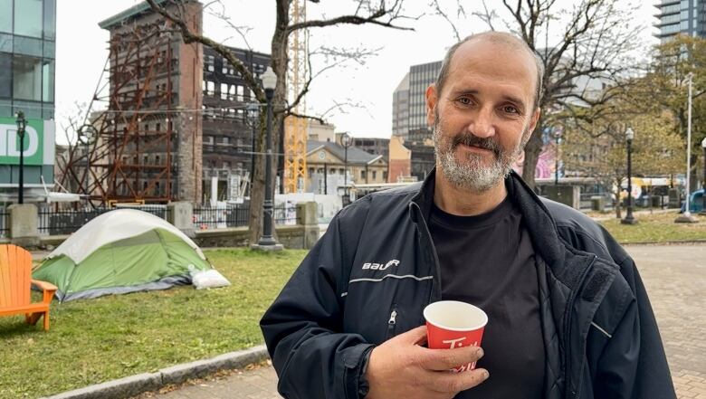 A man holding a cup of coffee stands in front of a tent in a public square in downtown Halifax.