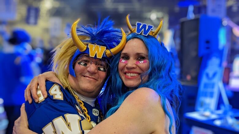 A man in a sports jersey with a horned helmet on, standing next to a woman in a blue wig.