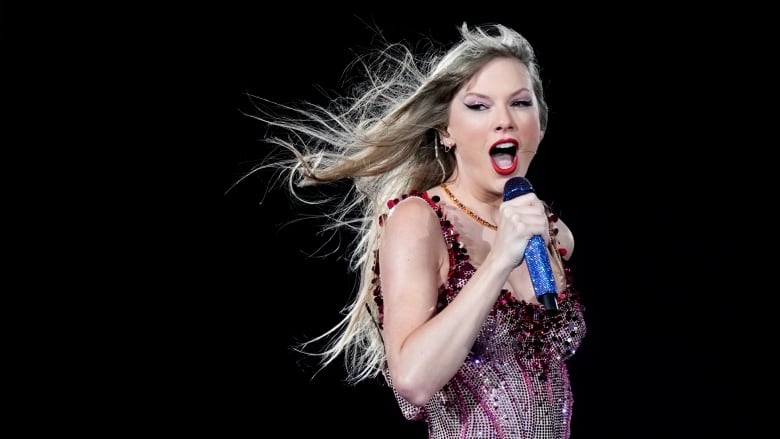 A women with long blonde hair blowing in a breeze sings into a blue-sparkled microphone.