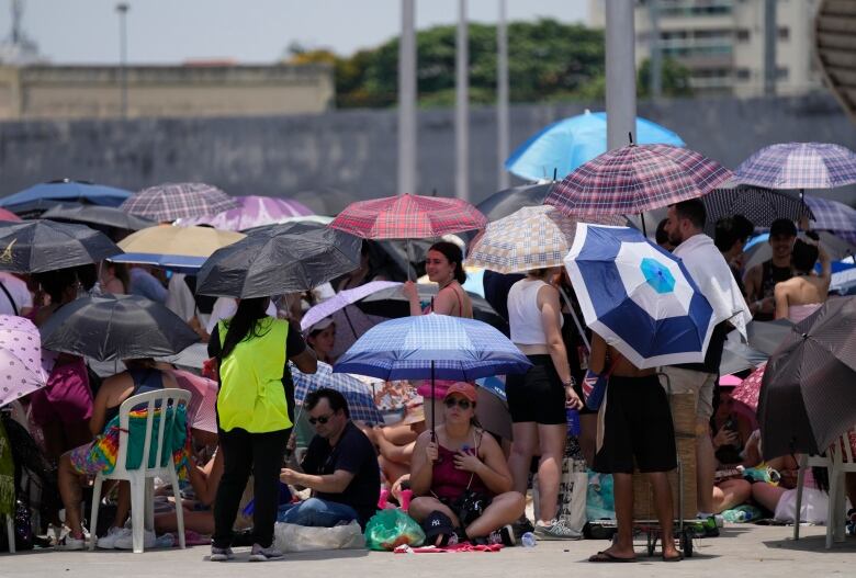 A crowd of people sit and stand underneath umbrellas, shielding themselves from the sun and heat.