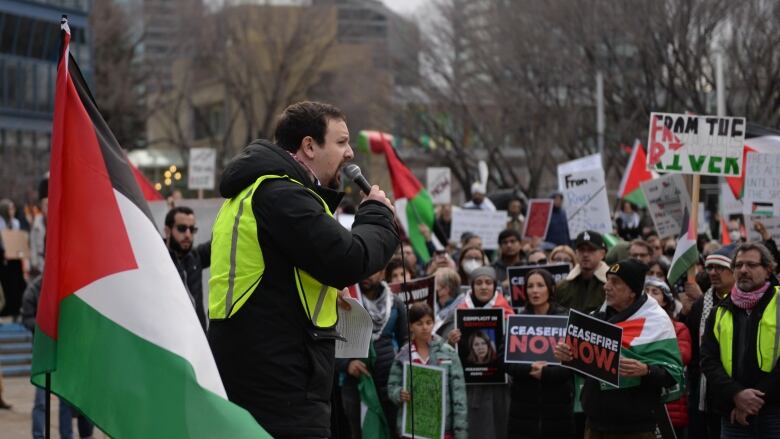 A man stands next to a Palestinian flag, speaking to a crowd at a protest.