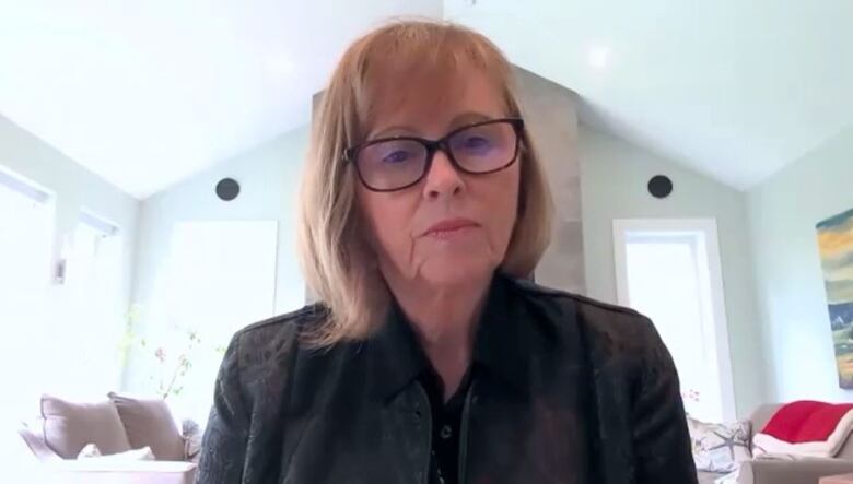 A white woman with glasses and shoulder-length reddish blonde hair sits at a desk with an open white living room behind her