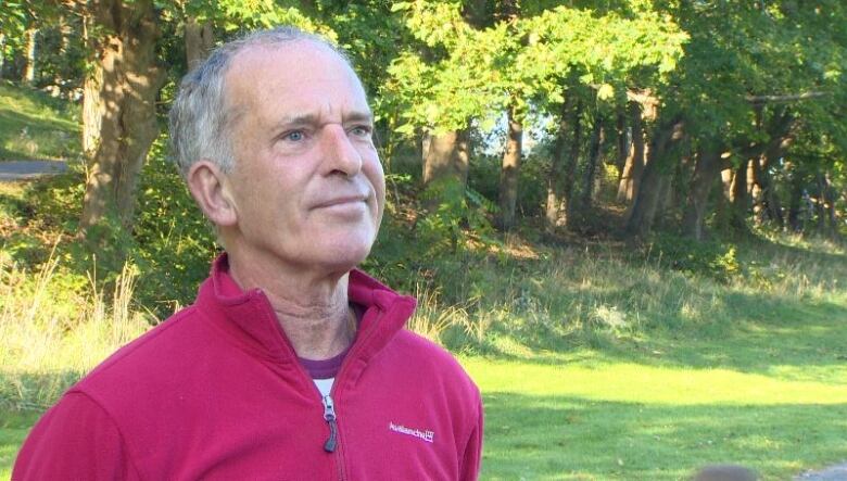 A white man in a red sweater stands in a park area with green trees behind him