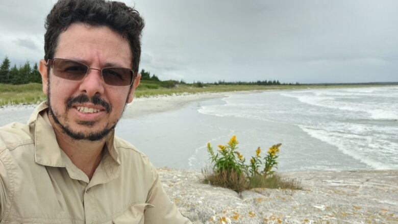 A man wearing sunglasses takes a selfie next to a beach.