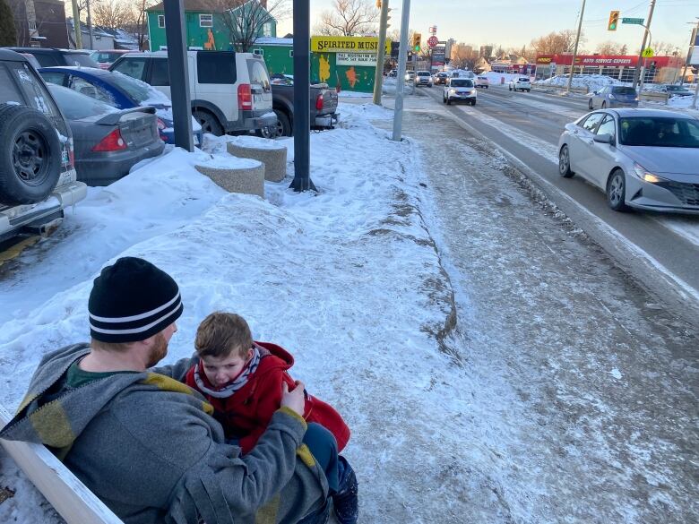 A man holds sits on a sidewalk bench holding a young child in winter conditions as vehicles move along at high speeds.