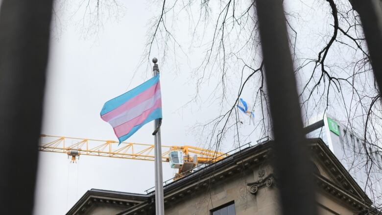 A transgender Pride flag is seen raised at Province House through the gates of the building. The Nova Scotia flag is to its right.