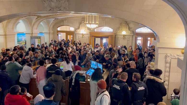 Protesters fill the Saskatchewan legislative building. Dozens of people can be seen around a security desk. Several peace officers are present.