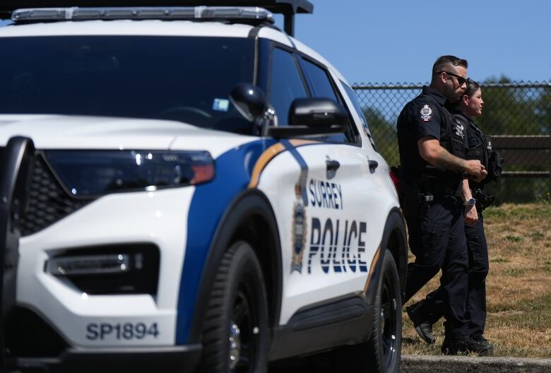 A blue and white police cruiser is pictured with Surrey Police written along the side. The car is facing forward in front of what looks like a baseball field and two uniformed officers (one male and one female) are visible walking behind the vehicle.