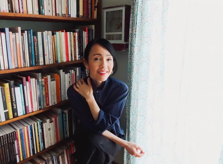 An Asian woman with short black hair poses in front of a bookshelf and a curtain. 