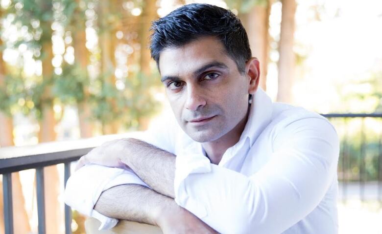 A brown man rests his arms on a chair and looks at the camera. There are trees in the background. 