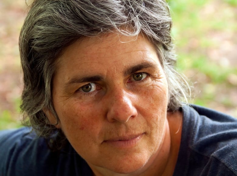 A white woman with grey hair looks at the camera. There is grass in the background and she wears a navy shirt.