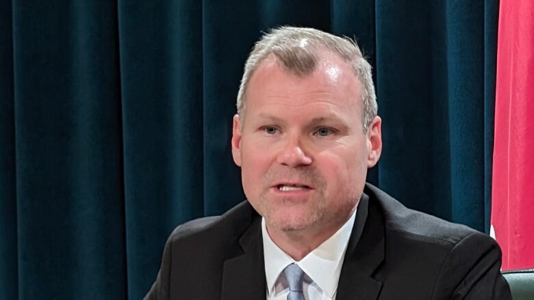 A white man with thinning grey hair is wearing a black suit jacket over a white dress shirt with a light blue tie. He is sitting in a leather desk chair, in front of some dark curtains and flags. 