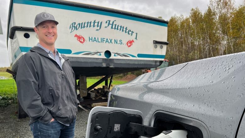 A white man in  rain jacket and jeans is seen wearing a ball cap. He is standing in front of his diesel boat and beside his truck while it uses electricity to charge.