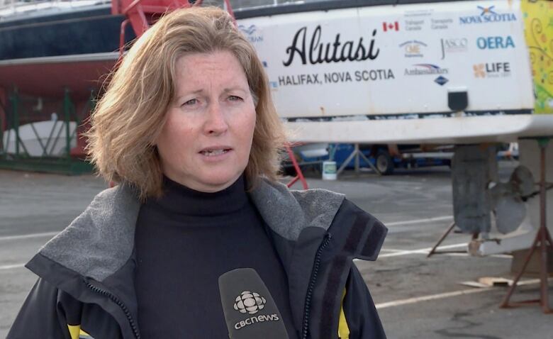 A white woman with strawberry-blonde hair in a bob stands in front of a mic. Behind her are boats hoisted onto lifts.