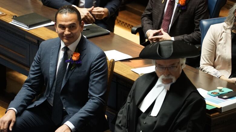 A man in a suit sits next to a man in robes and a tri-cornered hat in a legislative chamber.