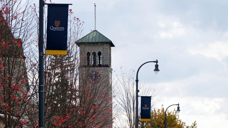 A limestone clocktower flanked by banners hanging from ornate light posts that have the coat of arms for Queen's University.