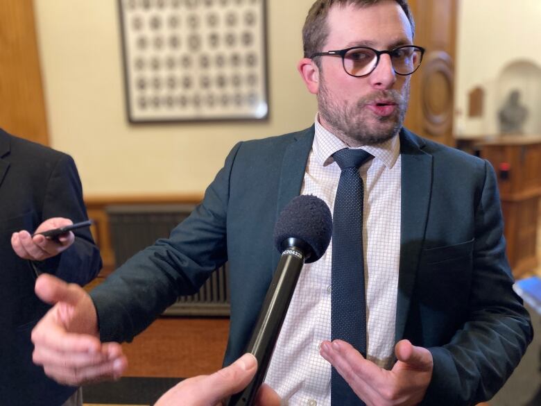 A young man with glasses in a suit gestures with his hands as he talks to reporters.