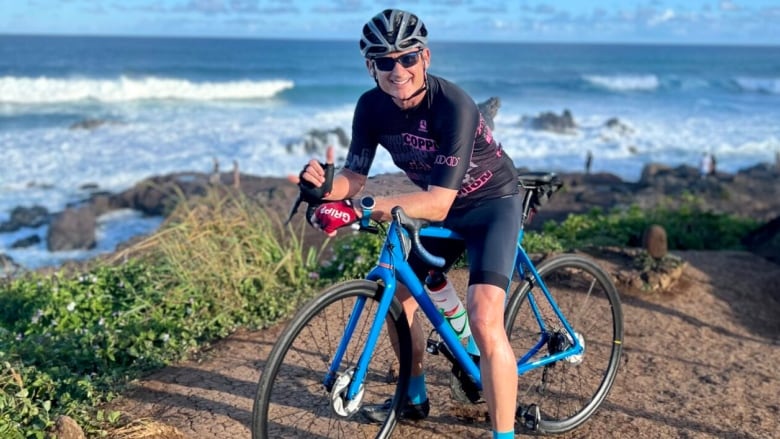 Mike Starko is shown posing in racing gear on a blue road bike on a cliff overlooking the ocean.