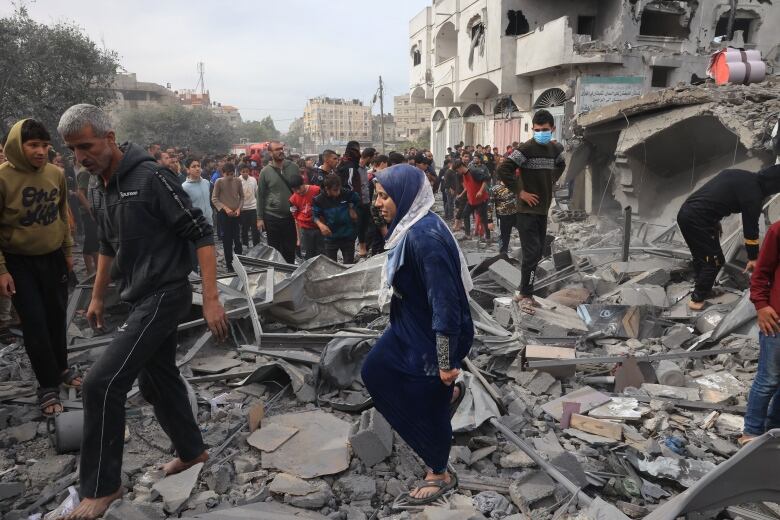 A man and a woman in a headscarf are shown in the foreground walking amid large amounts of rubble and concrete debris. A damaged building and several more people are shown in the background.