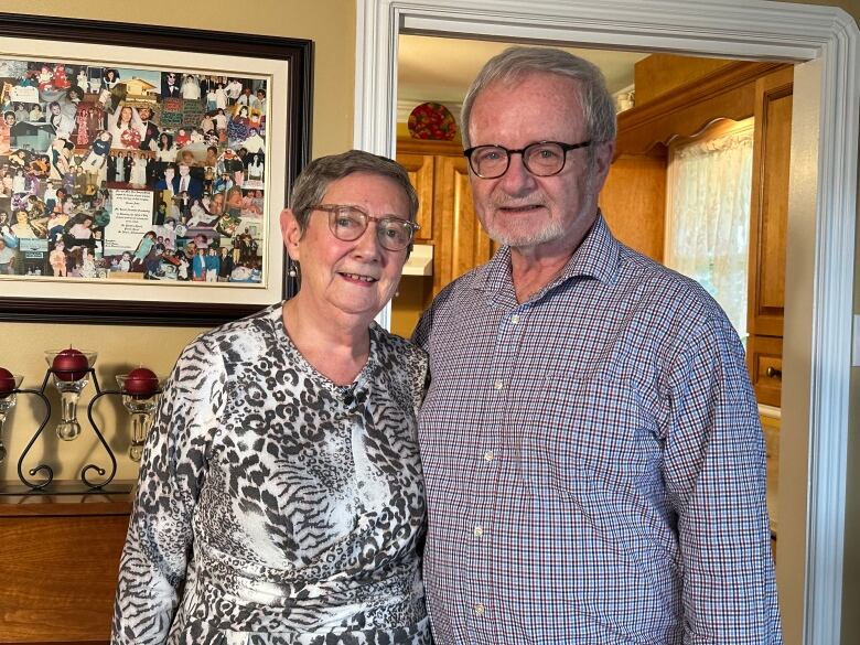 An elderly couple stand smiling in their living room.