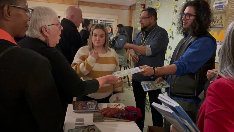 A group of people crowd around a table that has books on it. A man on one side of the table passes a book to a woman on the other side.