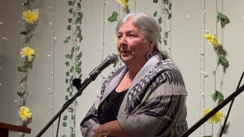 A woman with white hair pulled back in a ponytail wearing a grey, feather-patterned shawl stands in front of a microphone. 