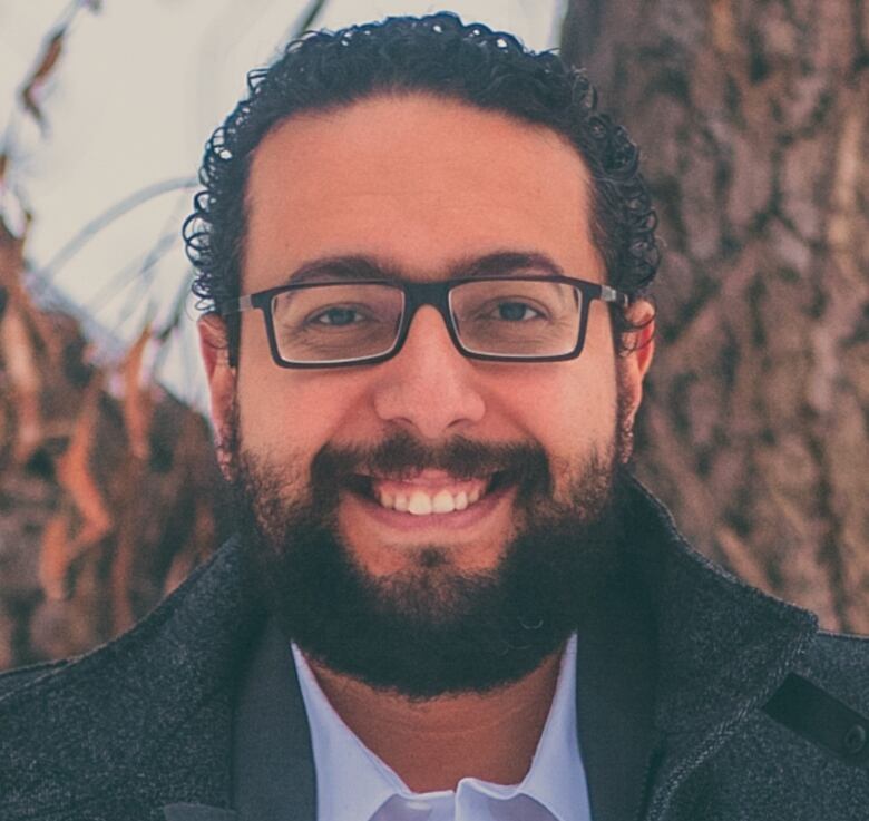 Ehab Diab is smiling with glasses and a suit jacket on, and there is a tree behind him