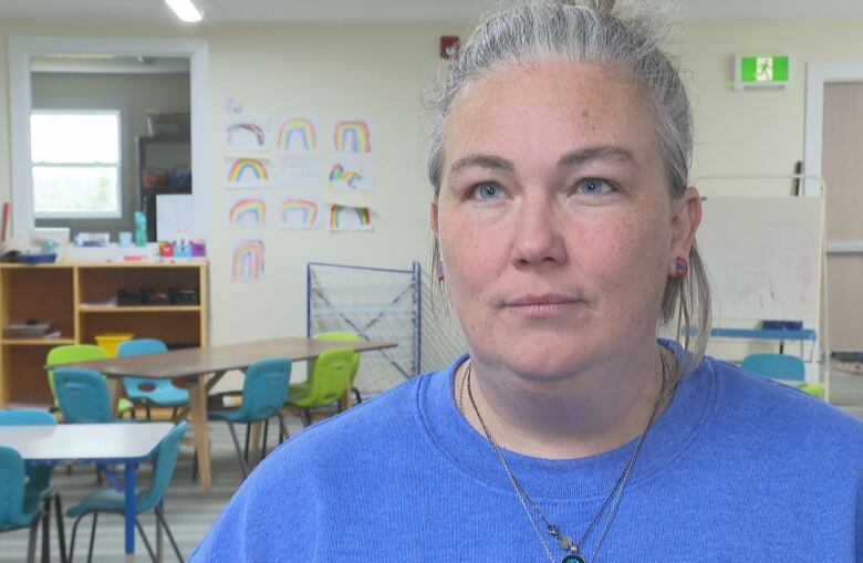 A person with grey hair tied back, wearing a blue sweater standing in front of small desks and chairs, and rainbow drawings on the wall.