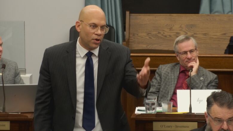 A man in a dark suit, white shirt and dark suit asking questions to the health minister.