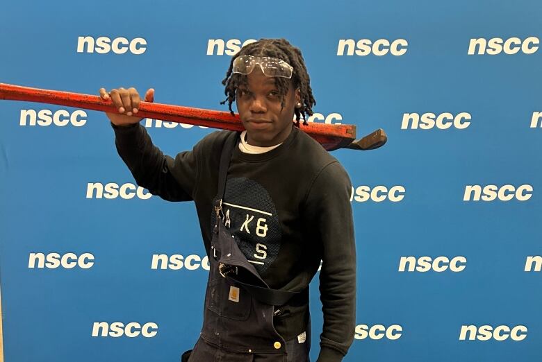 A man wearing overalls holds a wrench over his shoulder. he poses in front of a an NSCC backdrop. 