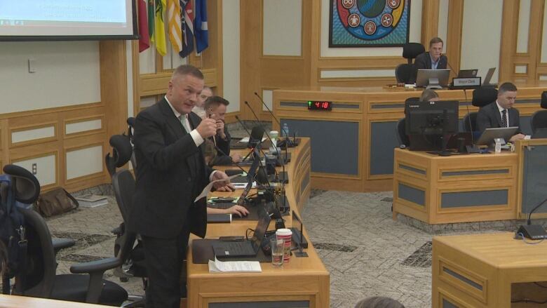 A man in a suit stands up from his chair in a city council chamber