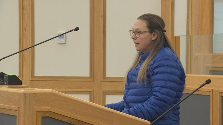 A woman in a puffy blue jacket speaks into a microphone at a podium